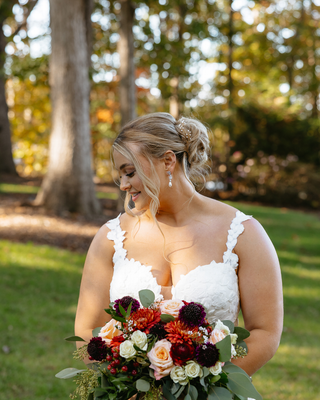 Bride in a white wedding dress looking down at a colorful bouquet of burgundy, coral, cream, and peach flowers with greenery.