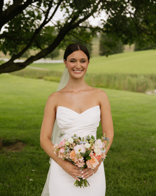 Bride in a strapless white wedding gown holding a pastel bouquet of pink, peach, and cream flowers outdoors.