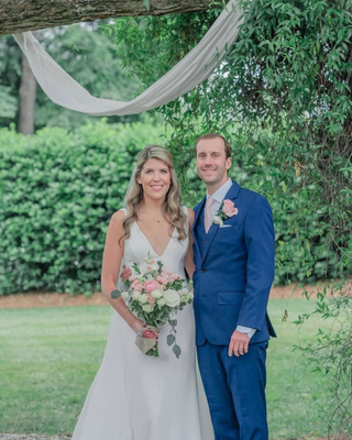 Bride and groom posing beneath a draped outdoor wedding canopy, holding a bouquet of blush pink and white flowers with eucalyptus greenery.