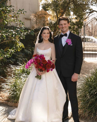 Bride and groom posing outdoors in a garden, holding a dramatic bouquet of burgundy roses, red flowers, and pink orchids.