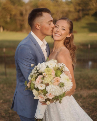 Groom kissing his bride on the cheek as they pose outdoors with a pastel bouquet of white, blush, and pale green flowers.