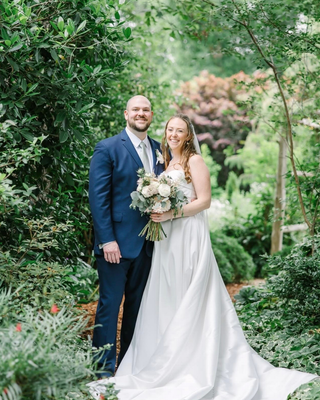 Bride and groom standing together in a garden surrounded by greenery, holding a white and blush bouquet with eucalyptus foliage.