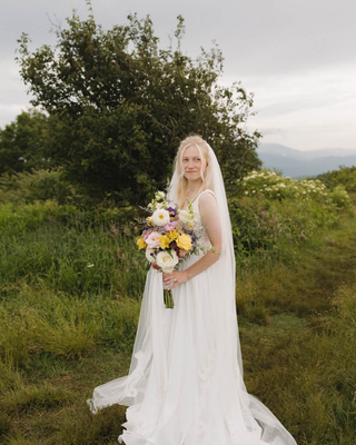 Bride in a white wedding gown standing in a grassy meadow overlook called The Beauty Spot with her colorful bridal bouquet.