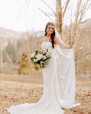Bride in a strapless white wedding gown holding a white and green bouquet while lifting her veil in a wooded setting.