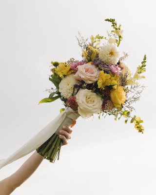 Bridal bouquet featuring yellow and blush roses, cream flowers, purple blooms, and greenery, wrapped with an ivory ribbon.