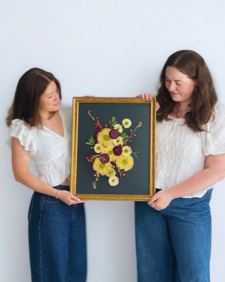 Two women holding pressed wedding flower art, in a vintage gold frame with black paper backing
