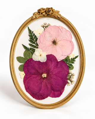 Pink and magenta pressed flowers with delicate green foliage arranged in a gold vintage oval frame.