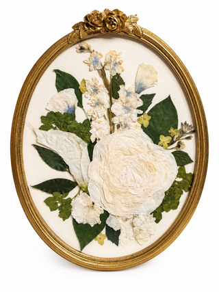 White pressed roses and small white flowers arranged with green foliage in an ornate gold vintage oval frame.
