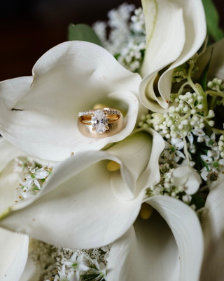 Gold diamond engagement ring resting on white calla lilies and wedding flowers.